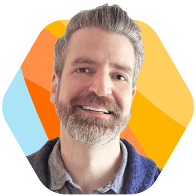 A smiling man with a grey beard and hair, wearing a button-down shirt and blue sweater, is framed within a rounded hexagon. The background features diagonal stripes in blue, orange, and yellow. This headshot is part of Bullhorn's Rally Cry - Run with the Bold campaign for the staffing industry.