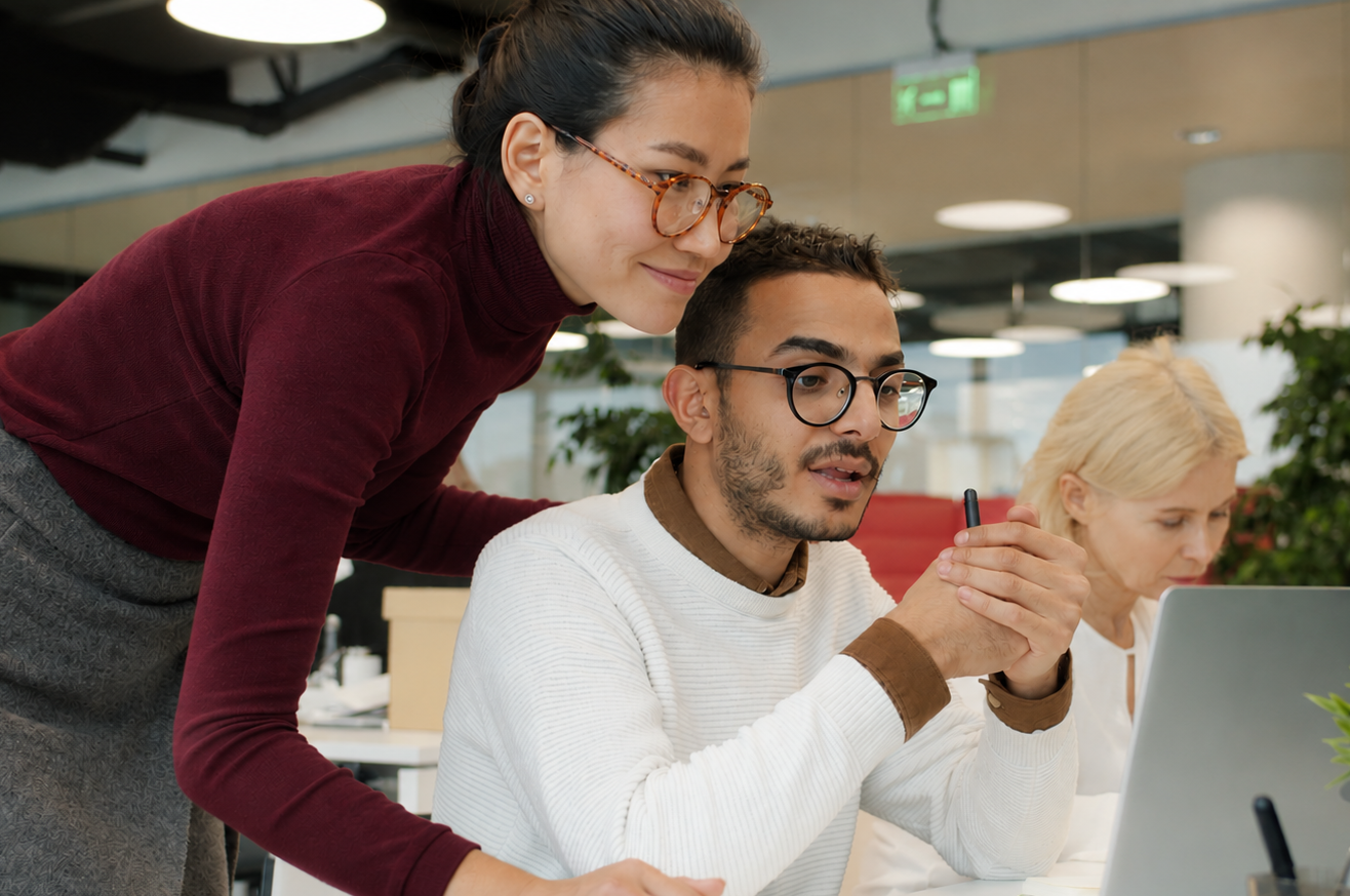 Coworkers collaborate around a laptop in a bright office, discussing work with focused expressions while a colleague works in the background.